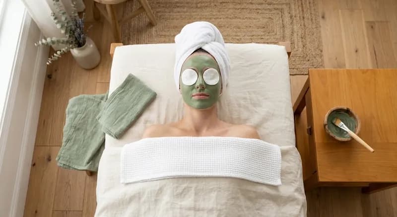 Tratamento de rosto a decorrer: mulher deitada numa marquesa com máscara de argila verde e os olhos tapados com discos de algodão